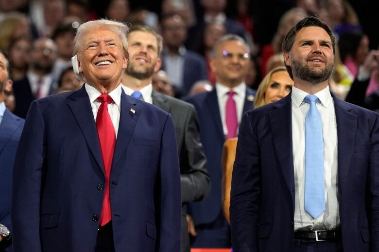 Donald Trump, in a dark blue suit and red tie, smiles with a bandage over his right ear. Next to him is a younger, bearded man in a dark blue suit and a light blue tie. A large crowd of people is in the background. 