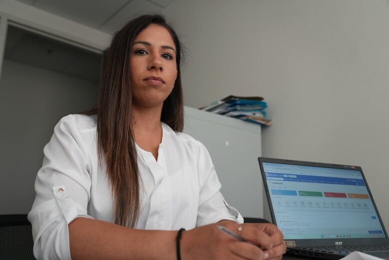 A woman with long, dark hair wearing a white shirt sits next to a computer screen. 