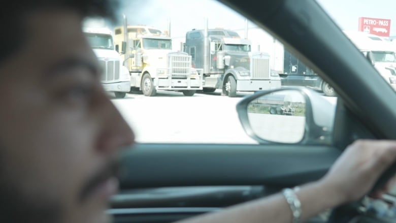 A man drives a car along a road where parked semi trucks are visible out the driver's side window.