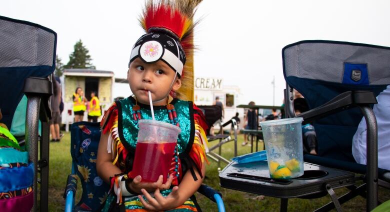 A small child in regalia holds a larger red drink, sipping from a straw.