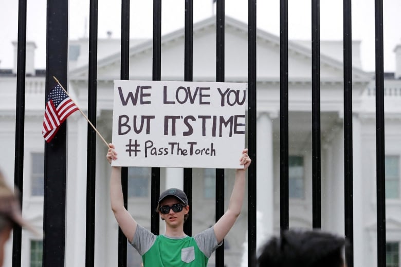 A protestor holds up a miniature American flag and a sign that reads, 'We love you but it's time #PassTheTorch.'