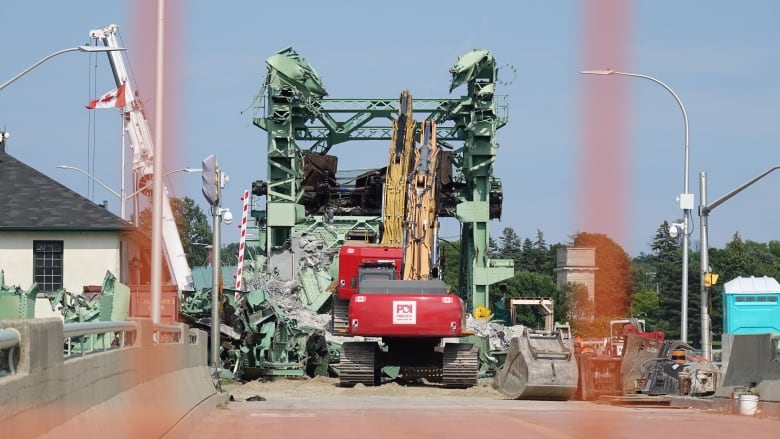 Pieces of heavy equipment can be seen parked in front of a twisted green, metal bridge and piles of rubble.