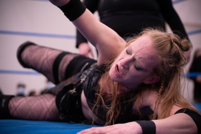 Female wrestler with long, red hair lays on mat with pained look on her face. Her right arm is stretched up off her body. A person in black stands behind her. 