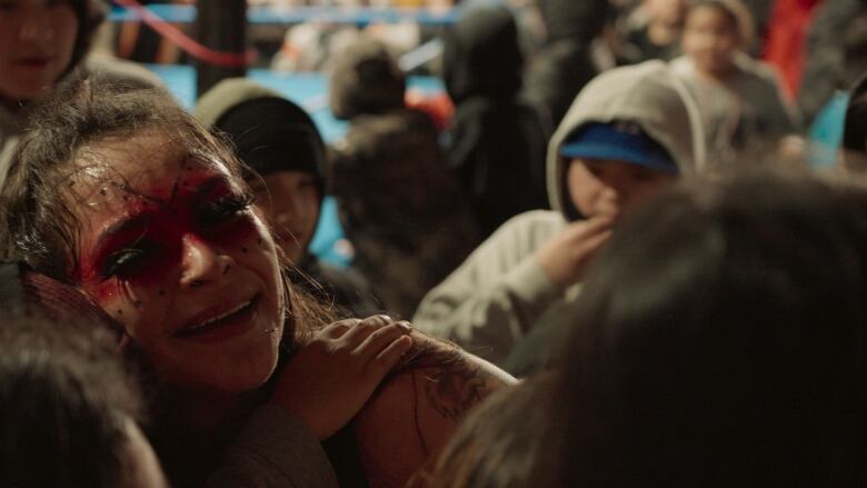 Closeup on face of female wrestler as her gets hugs from youth. Empty wrestling ring in the background with a youth wearing a hoodie in front of it. 