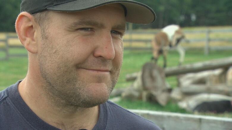 A young man in a ball cap stands outside speaking in front of a goat blurred in the background.