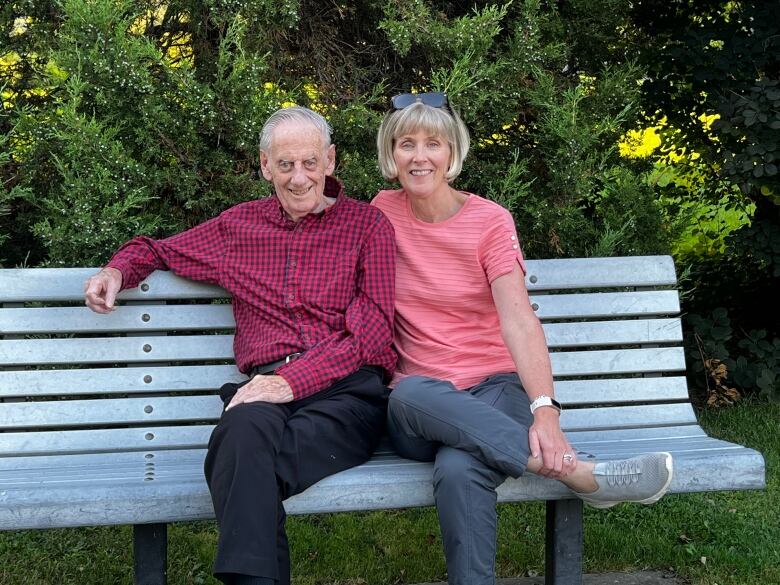 An elderly man in a red plaid shirt sits next to a younger woman with a blonde pixie cut and a pink shirt on a bench.