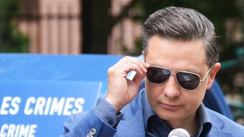 A man in a blue blazer lifts his hand to remove a pair of aviator sunglasses.