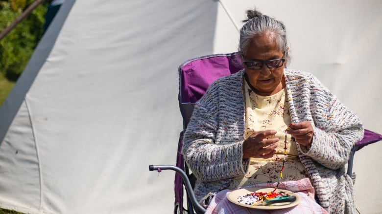 A woman sits in front of a teepee making a beaded necklace.