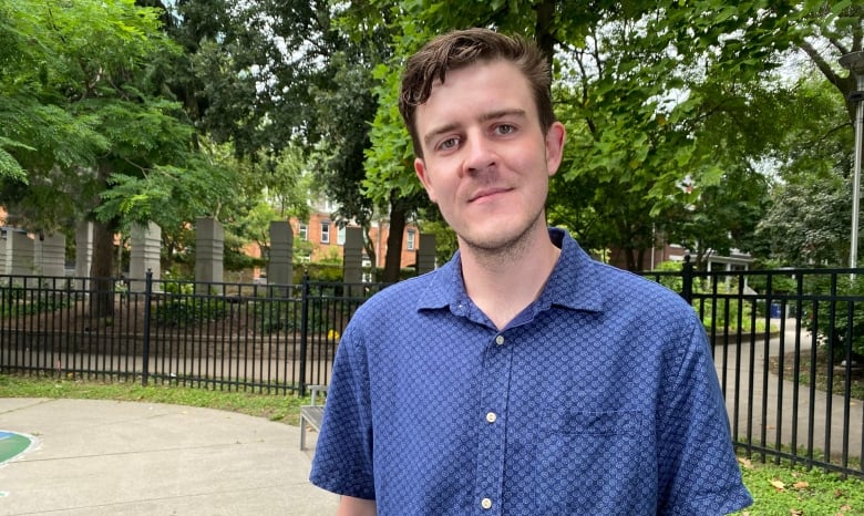 Curran Stikuts, wearing a blue collared shirt, standing outdoors with a fence and trees behind him. 