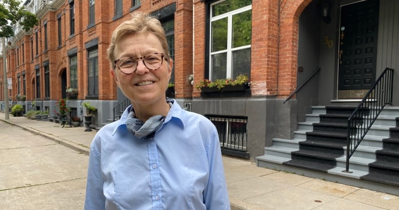 Vera Tarman, wearing glasses and a blue collared shirt, stands in front of a row of brick houses. 