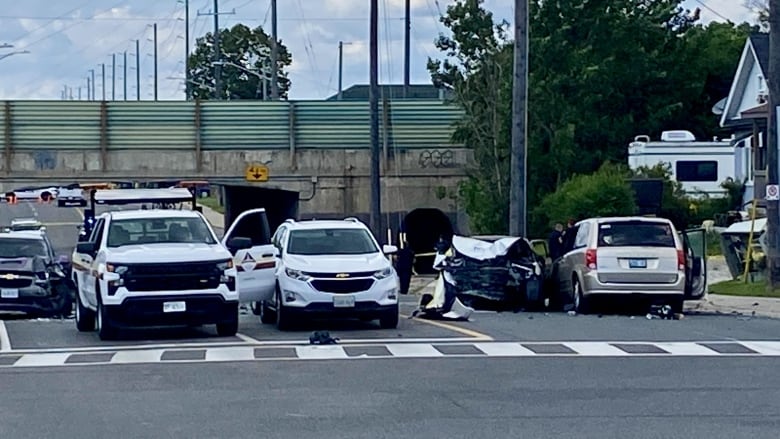 Cars are stopped on a 4-lane city street in broad daylight. There's an overpass close in the distance. To the right, against the curb, a mini van is stopped against a smashed vehicle facing the opposite direction