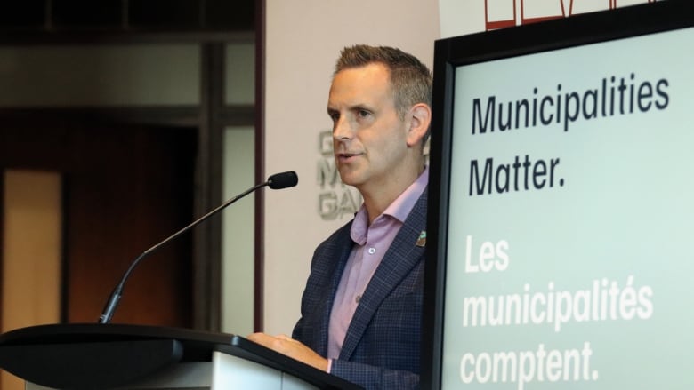 A man in a suit jacket speaking into a microphone with a TV showing 