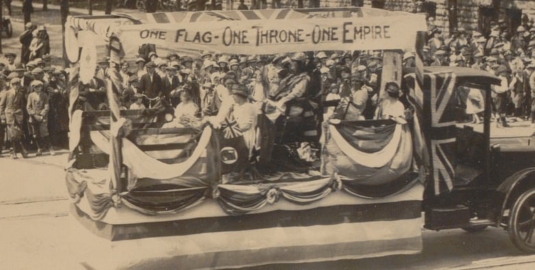 Black and white photo of a truck with people in the back and decorations for the British Empire.