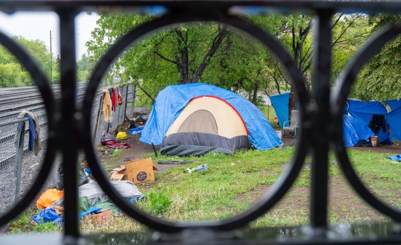 A cluster of tents is seen in a grassy area next to train tracks, through a black fence.