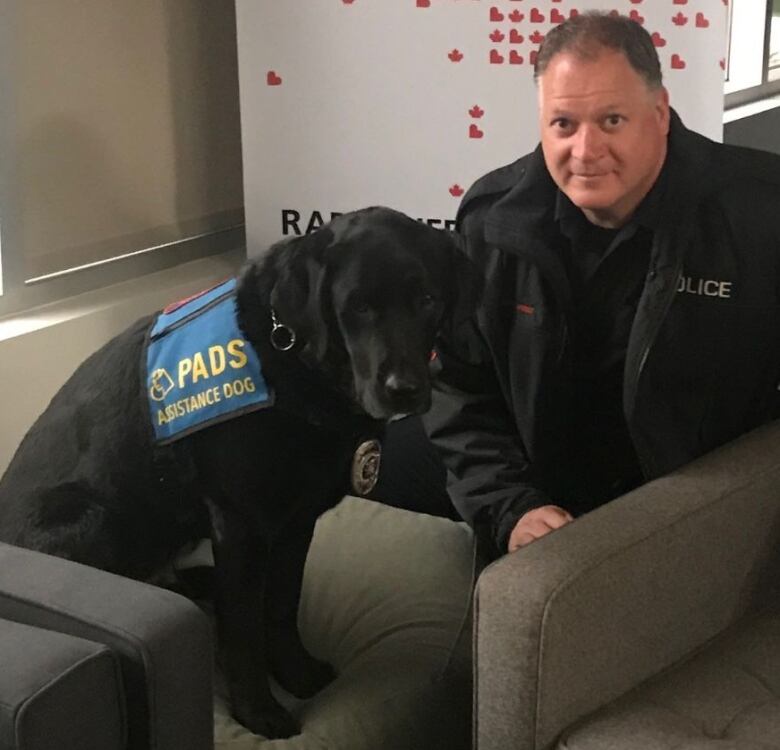 a man in a police uniform poses next to a black service dog.