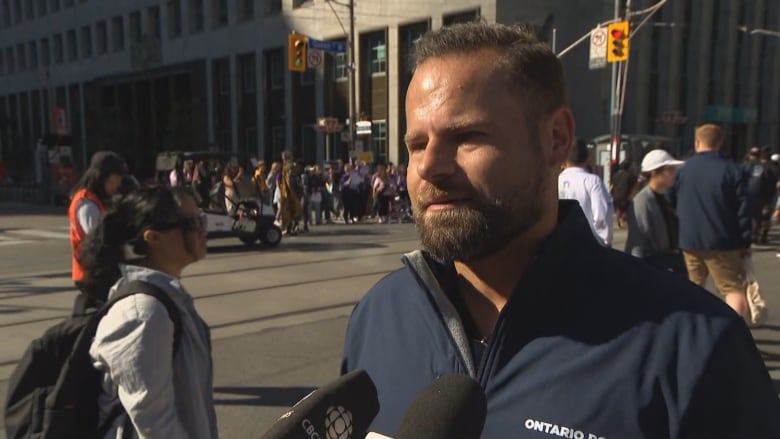 A bearded man in a blue shirt stands in front of a busy city street on a sunny day. There are CBC microphones in front of him