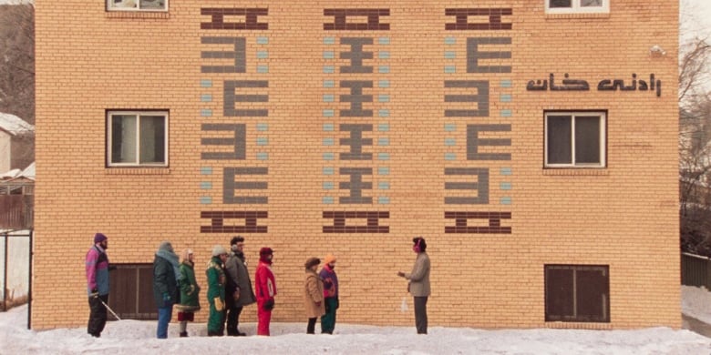 Several people standing in a line on a snowy sidewalk outside a brick apartment building.