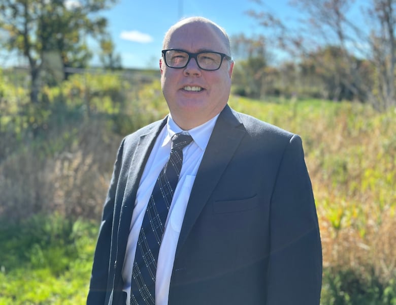 A person wearing a suit and tie stands in front of a grassy field.