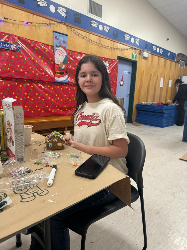 A woman sits at a table with a gingerbread house. 